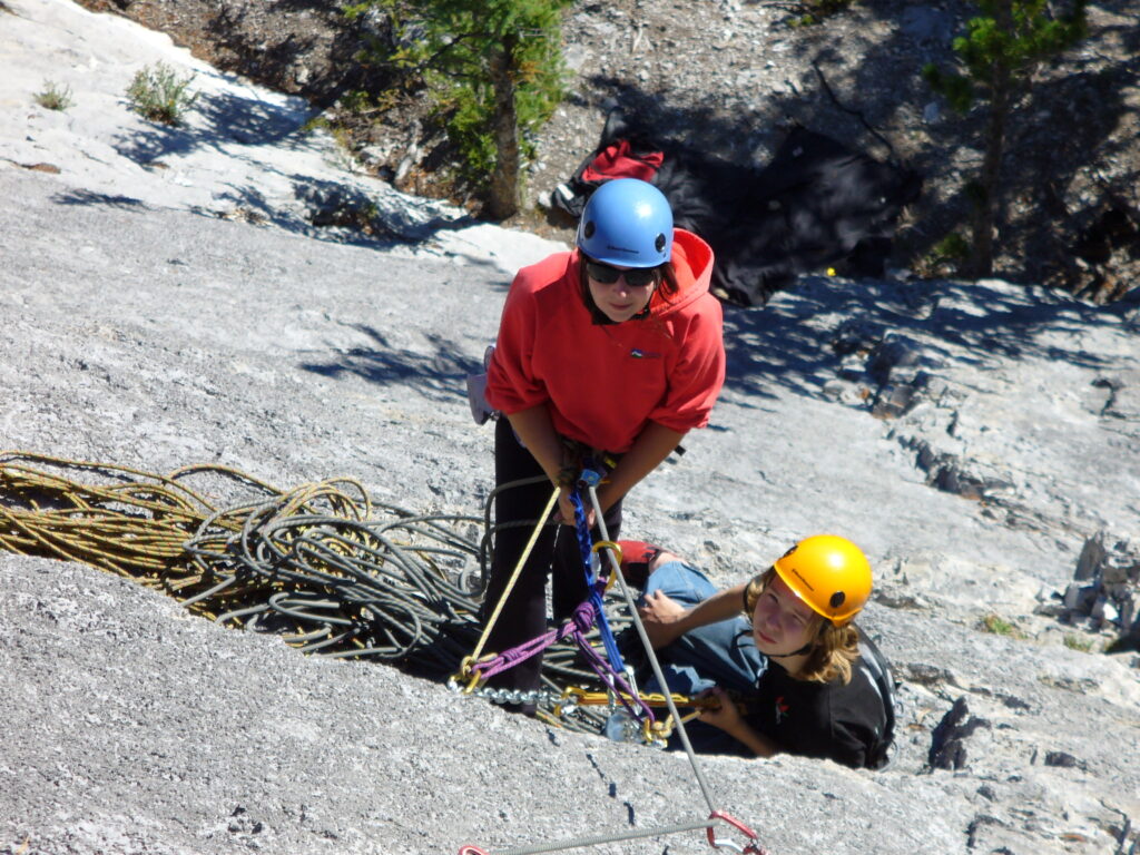 Looking down at two people on a very thin ledge half way up a sheer mountain side, tied into  an anchor point. A coil of rope is at their feet.