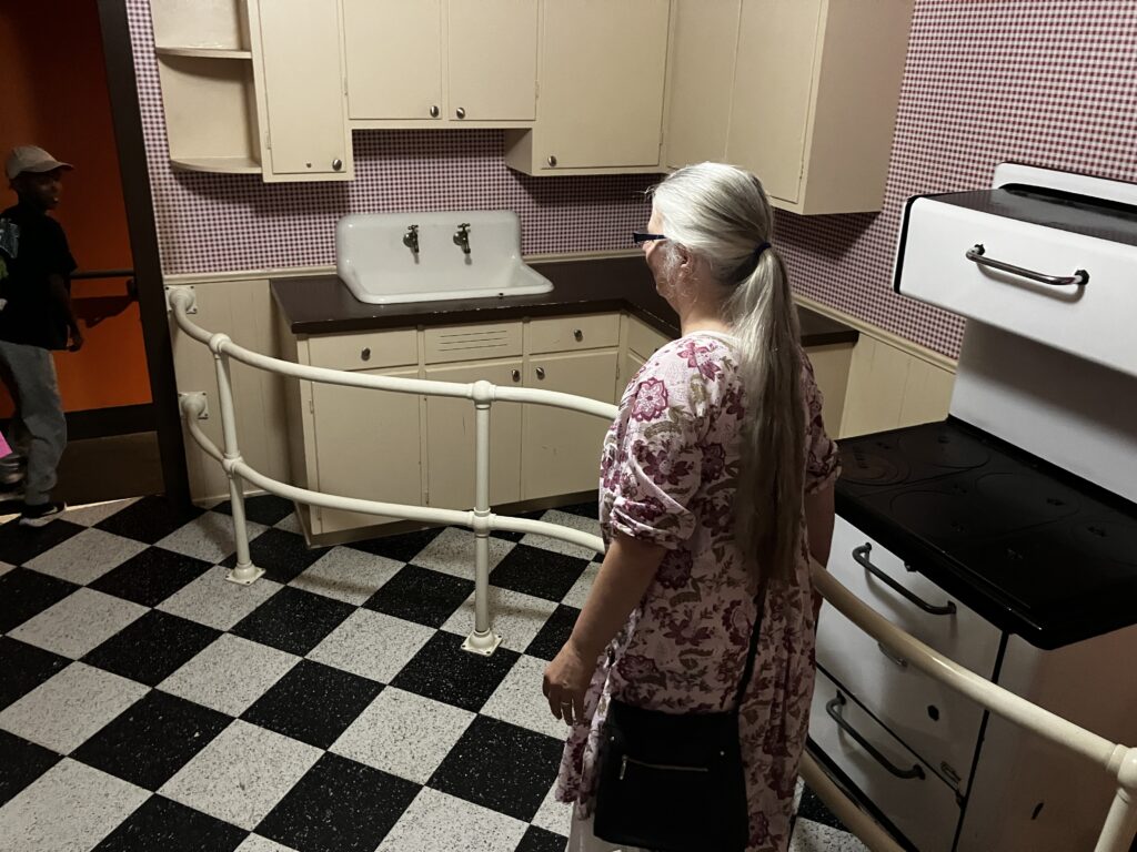 A person stands in an old fashioned kitchen with a black and white checked floor. What si not obvious is the whole kitchen is tilted